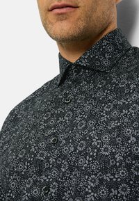 Schwarzes Hemd mit floralem Muster, das einen Kragen, einen Knopfverschluss und einen strukturierten Stoff aufweist. Die Muster bestehen aus kleinen Blumen und Blättern.