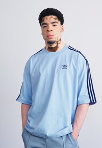 Šviesiai mėlynas medvilninis marškinėliai trumpomis rankovėmis, su tamsiai mėlynomis juostelėmis ant pečių ir mažu juodu Adidas logotipu ant krūtinės.