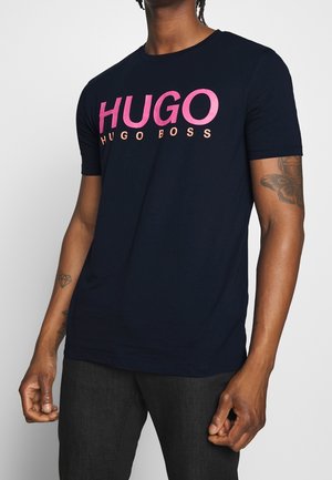 Osoba oblečená v černém tričku s krátkým rukávem s růžovým logem "HUGO BOSS", viditelné tetování na levém předloktí a černé kalhoty.