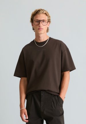 Jeune homme aux cheveux blonds bouclés portant de grandes lunettes rondes, un t-shirt noir oversize, un pantalon noir, un collier chaîne argenté et un bracelet, debout avec une main dans la poche.