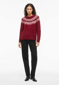 Femme portant un pull en tricot rouge à motifs, un pantalon noir à jambe droite et des bottes de cheville à talons noires, se tenant devant un fond blanc.