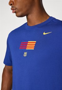 Blå bomulds t-shirt med rund hals. Har et grafisk design med røde og gule striber samt et skjoldlogo nedenfor. Nike-logo i gult på skulderen.