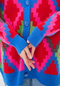 Gebreide cardigan met een levendig patroon in roze, rood en groen, met een blauwe knoopsluiting aan de voorkant en geribde manchetten. De textuur ziet er zacht uit.