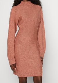 Robe en tricot couleur rouille avec un col montant, des manches longues et une texture douce, dotée d'une silhouette cintrée et de poignets à côtes.