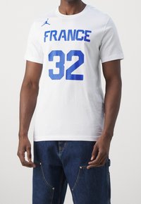 Biała bawełniana koszulka z niebieskim napisem „FRANCE” i numerem „32”. Posiada okrągły dekolt i krótkie rękawy. Logo Jordan na ramieniu.