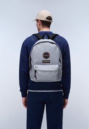 Man wearing navy tracksuit and beige cap, carrying light gray backpack with front pocket and round black logo patch, facing away on plain background.