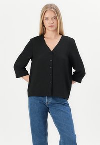 Svart v-hals bluse med trekvart ermer, med strukturert stoff og seks svarte knapper foran. Kombinert med blå jeans.