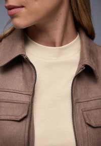 Gros plan sur le bas du visage, le cou et le haut de la poitrine d'une personne portant une chemise beige sous une veste marron partiellement zippée.