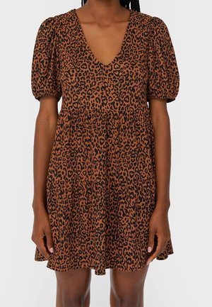 Femme portant une robe courte marron avec imprimé léopard noir, manches courtes bouffantes et col en V, debout, les mains relâchées le long du corps.