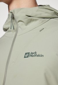 Giacca leggera di un verde chiaro, con colletto alto, chiusura a zip e logo "Jack Wolfskin" in verde scuro sul petto. Tocco morbido.