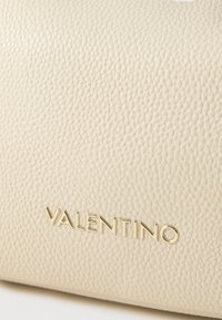 Beige handtas van getextureerd leer met een goudkleurig "VALENTINO" logo dat duidelijk zichtbaar aan de voorkant is geplaatst.