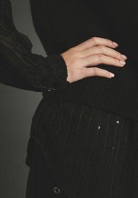 Pull noir avec texture côtelée, manches transparentes noires rayées avec accents de pois et ourlet incurvé. Main reposant sur le vêtement.