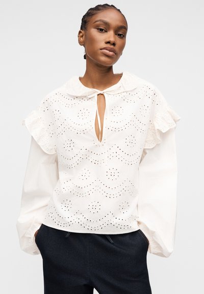 Femme aux cheveux tressés portant un chemisier blanc à œillets avec des épaules à volants et des manches longues, associée à un pantalon foncé, debout les mains dans les poches.