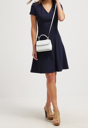 Vrouw die een marineblauwe V-hals jurk met korte mouwen draagt, beige open-toe hakken en een kleine witte crossbody handtas met een bovenhandgreep.