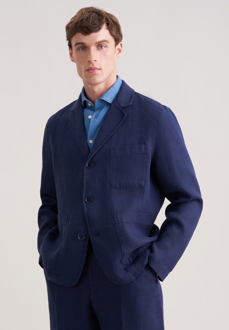 Jeune homme portant une veste bleu marine par-dessus une chemise à boutons bleu clair, debout les mains dans les poches devant un fond uni.