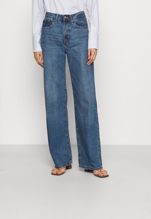 Persoon draagt een blauwe spijkerbroek met hoge taille en brede pijpen, een wit shirt met lange mouwen ingestopt en open-toe sandalen met hak, staand tegen een neutrale achtergrond.