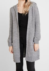 Grå stickad kofta med huva, med ett strukturerat mönster, ribbade ärmslut och sidofickor. Bärs över en svart topp och byxor.