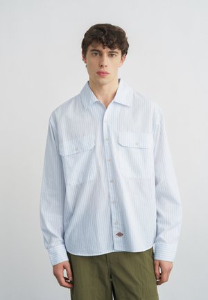 Junger Mann trägt ein hellblau gestreiftes Hemd mit Knöpfen und zwei Brusttaschen sowie olivgrüne Hose, steht vor einem einfarbigen Hintergrund.