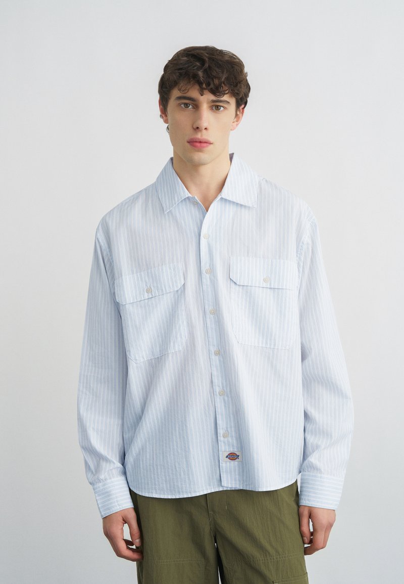 Νέος άνδρας που φοράει ανοιχτό μπλε πουκάμισο με ρίγες και δύο μπροστινές τσέπες, καθώς και παντελόνι ελιάς, στέκεται μπροστά σε απλό φόντο.