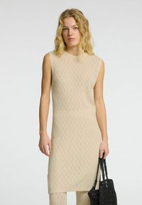 Robe en tricot beige avec un motif en vagues, sans manches, et un col à côtes. Le mannequin tient un sac fourre-tout en crochet noir.