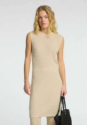 Beiges Strickkleid mit Wellenmuster, ärmellosem Design und geripptem Ausschnitt. Das Model hält eine schwarze gehäkelte Tragetasche.