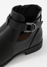 Bottine noire en cuir lisse avec un motif texturé effet crocodile. Une boucle métallique orne le côté. Design à petit talon plat.