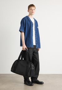 Svart duffelbag med doble håndtak, laget av slitesterkt stoff. Har en glatt tekstur og subtile detaljer, holdt av en person i en denimskjorte.