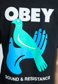 Must t-särk mustast puuvillast, millel on graafika türkiissinisest lindudest, kes istuvad sinisel käel, ja tekst "KUULA JA RESISTEERI" valges.