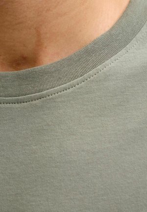 Približnji posnetek vratu in zgornjega dela prsnega koša osebe, ki nosi svetlo zeleno majico z okroglim izrezom in vidnim šivanjem.