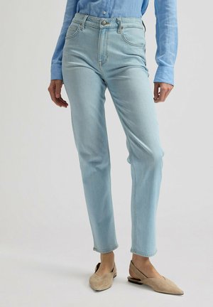 Osoba ubrana w jasnoniebieskie dżinsy, niebieską koszulę na guziki oraz beżowe baleriny z szpiczastym noskiem, stojąca na tle jednolitego tła.