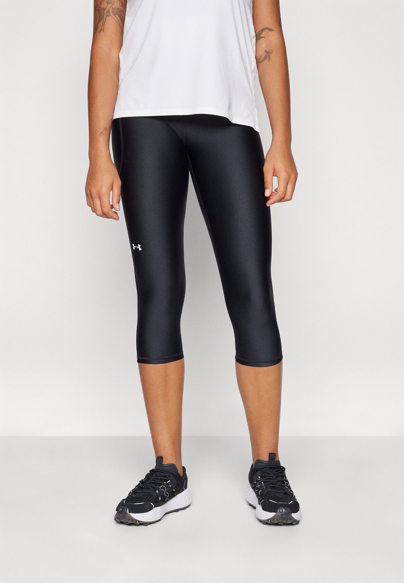 Personne portant un legging court noir Under Armour, un haut sans manches blanc, des chaussures de sport noires et blanches, debout devant un fond uni.