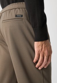 Smeđi, mekani hlače od tkanine s bočnom džepom i crnom oznakom Calvin Klein, prikazane uz teksturiranu crnu rukavicu i ruku.