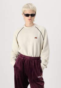 Cremefarvet sweatshirt med lange ærmer, sort kantbånd og et lille rødt og orange logo. Matchet med bordeaux velour joggers med hvid logo tekst.