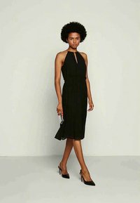 Γυναίκα με αφρο χτένισμα, φορώντας ένα μαύρο μέσο φόρεμα χωρίς μανίκια και μαύρες γόβες, κρατώντας μία μικρή ασημένια τσάντα, που στέκεται απέναντι από ένα απλό φόντο.