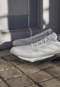 Valkoiset Adidas-juoksukengät, joissa on rei'itetty päällinen ja "Dreamstrike+"-teksti mustassa pohjassa, asetettu kiviselle laatoitetulle pinnalle lähellä harmaata seinää.