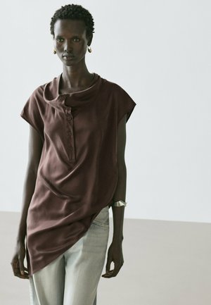 Femme aux cheveux courts et bouclés portant un débardeur marron ample et un jean bleu clair, debout devant un fond uni.