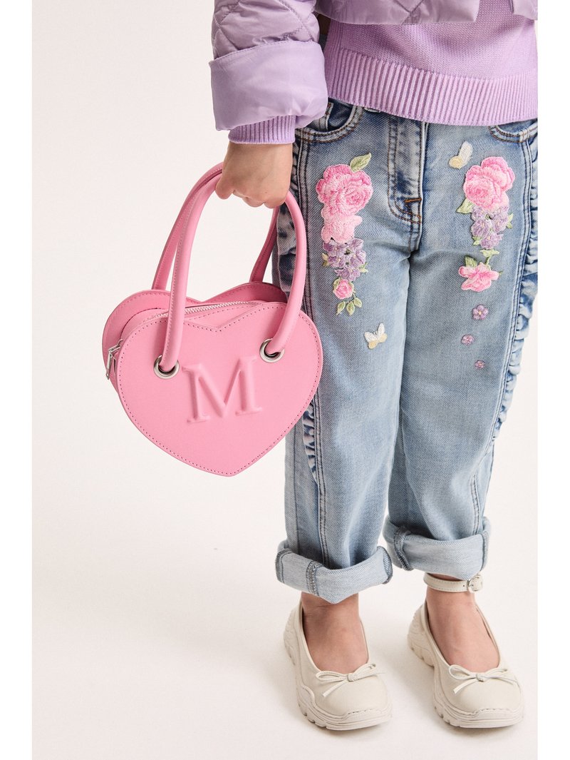 Παιδί που φοράει τζιν με κεντητά λουλούδια και λιλά πουλόβερ, κρατώντας μια ροζ τσάντα σε σχήμα καρδιάς με ανάγλυφο «M».