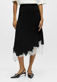 Naine kannab musta midi seelikut, mille ääres on valgete pitsdetailidega ääristus, koos mustade madalate kontsadega terava ninaga slingback kingadega.