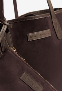 Bruine suède schoudertas met een gladde afwerking, voorzien van een leren merklabel en een bijpassend klein ritsvakje eraan bevestigd.