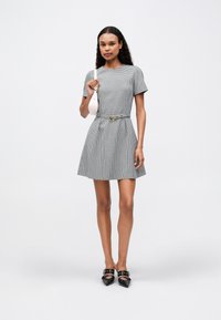 Γυναίκα ντυμένη με ένα καρό φόρεμα κοντού μανικιού με ζώνη, κρατώντας μια μικρή λευκή τσάντα ώμου και μαύρες γόβες με λουριά.