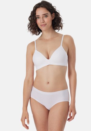 Kobieta o kręconych włosach, ubrana w biały biustonosz i dopasowaną bezszwową bieliznę, stojąca na tle jednolitego jasnego tła, lekko się uśmiechająca.