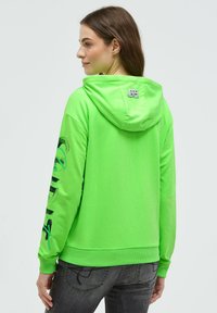 Leuchtend grüner Hoodie aus weichem Stoff, mit Kordelzug-Kapuze und Grafikdruck entlang des linken Ärmels. Gerippte Bündchen und Saum.