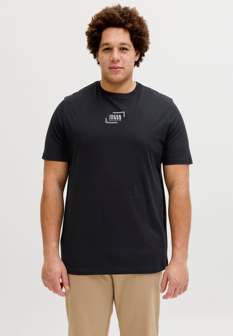 Άνδρας με σγουρά μαλλιά που φοράει μαύρο T-shirt Jack & Jones και μπεζ παντελόνι, στέκεται μπροστά σε απλό φωτεινό φόντο.