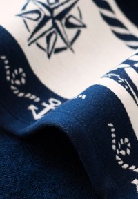 Granatowy materiał z białymi wzorami marynistycznymi, w tym kompasem i kotwicami, zwisający i częściowo złożony, ukazujący miękką teksturę.