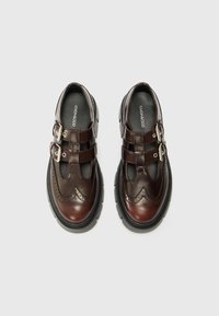 Scarpe in pelle marrone con design brogue, punta chiusa, cinturini doppi a fibbia e suola spessa in gomma nera. Texture liscia e forma classica.