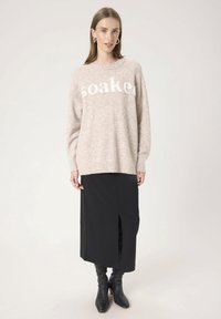 Femme portant un pull beige « mouillé », une jupe midi noire avec fente avant et des bottes à talons noires, se tenant devant un fond clair.