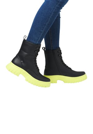 Black leather ankle boots feature a chunky neon yellow sole, with lace and side zipper closures for ease of wear.
