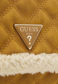 Tkanina z pikowanej musztardowej zamszu z metalowym, trójkątnym logo Guess U.S.A. oraz poziomym paskiem białego puszystego barana.