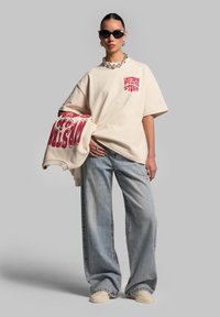 Bežové oversized tričko s červenou grafikou, spárované se světle modrými širokými džíny a bežovými slip-on botami. Modelka nosí černé sluneční brýle.