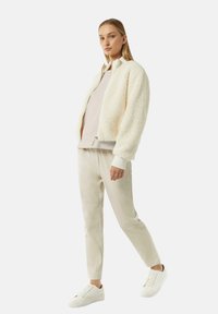 Cream-colored faux fur bomber jacket with a zip front, ribbed cuffs, and a relaxed fit, paired with beige pants and white sneakers.